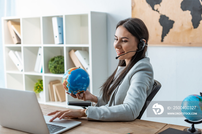 positive travel agent holding globe while sitting at workplace