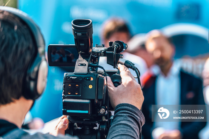 Cameraman At An Outdoor Media Event. Blurred Speaker In The Background