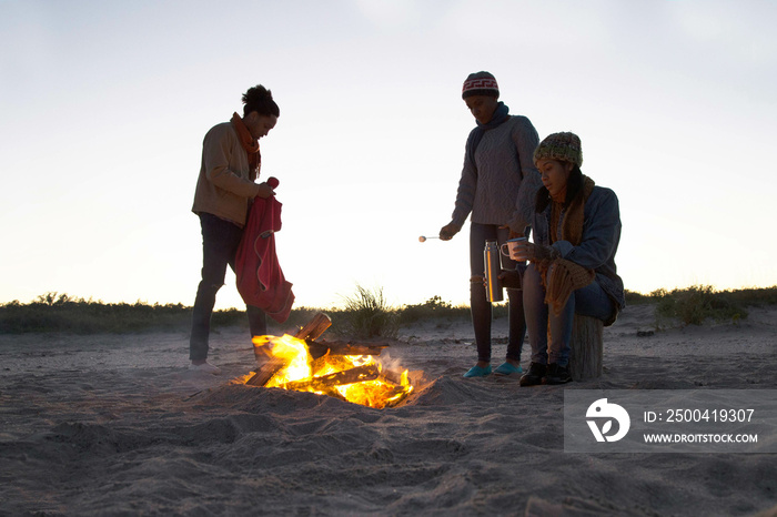 Three friends on beach, gathered around camp fire