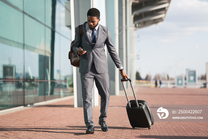 African Entrepreneur Using Phone Walking With Travel Suitcase At Airport