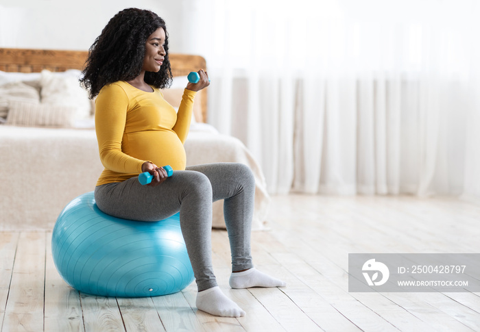 Active pregnant lady exercising with barbells while sitting on ball