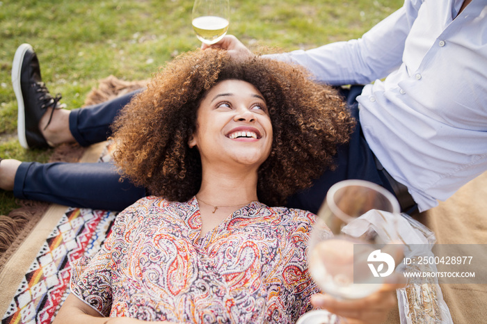 High angle view of happy girlfriend with wineglass talking while relaxing on boyfriends lap in park