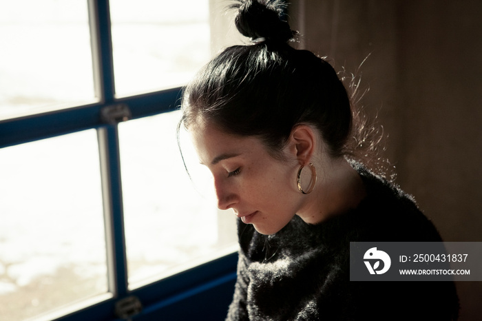 Close-up of thoughtful woman looking down while sitting against window at home