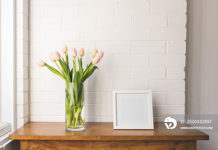 Close up of pale pink tulips in glass vase with blank square frame on wooden oak table against white