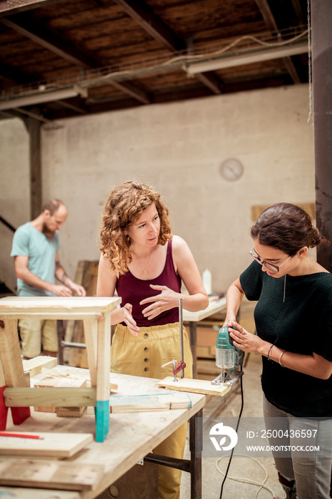 female carpenter guiding colleague cutting wood with jigsaw at workbench in workshop