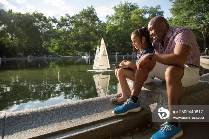 Father and daughter (6-7) sitting by pond in park, looking at view surfboards in background