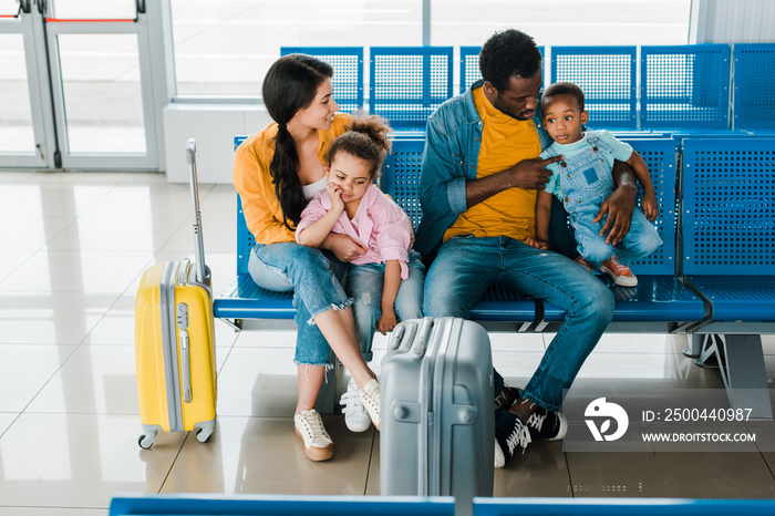 cheerful african american family with travel bags and kids sitting in airport in waiting hall