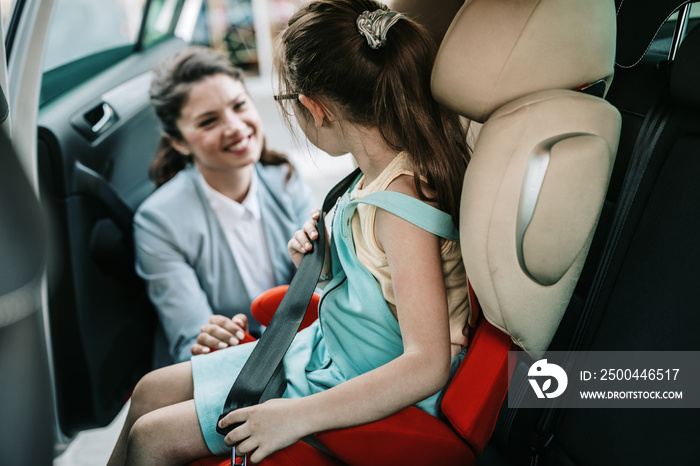 Young businesswoman helping her daughter to fasten seatbelts in the car while girl is sitting on a s