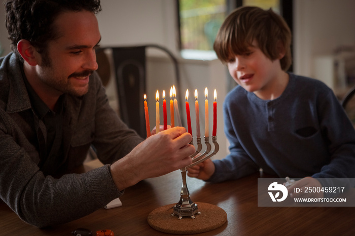 Boy looking at smiling father burning candles on menorah during Hanukkah festival