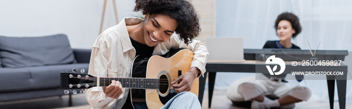 Young african american man playing acoustic guitar near blurred girlfriend in living room, banner.