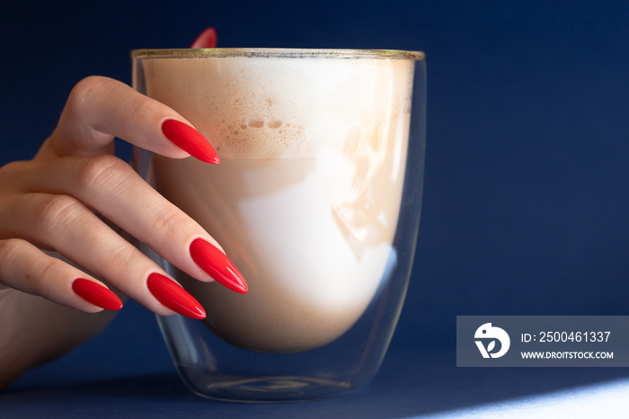 Girl`s hand with a beautiful red manicured nails and glass of coffee on the blue background