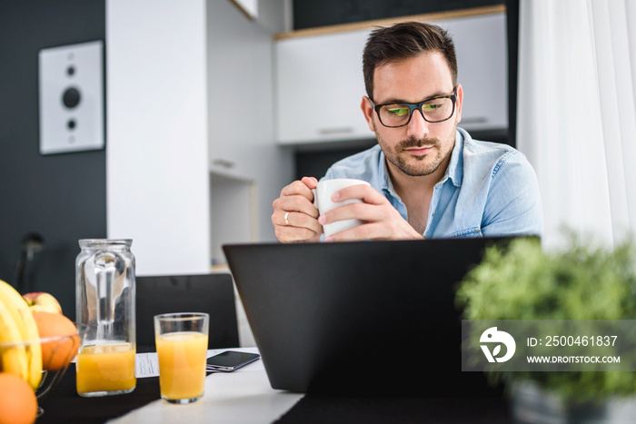 Handsome man using laptop computer at home, drinking juice and coffee in the morning. Working From H