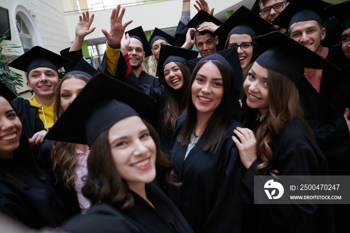 group of happy international students in mortar boards and bachelor gowns with diplomas taking selfi