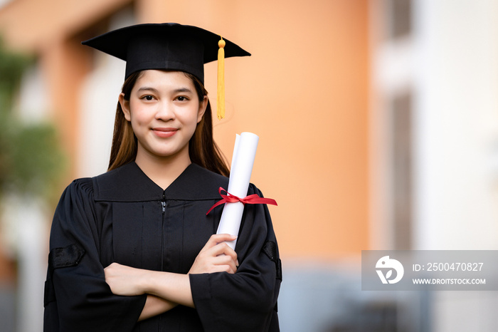 A young happy Asian woman university graduate in graduation gown and mortarboard holds a degree cert