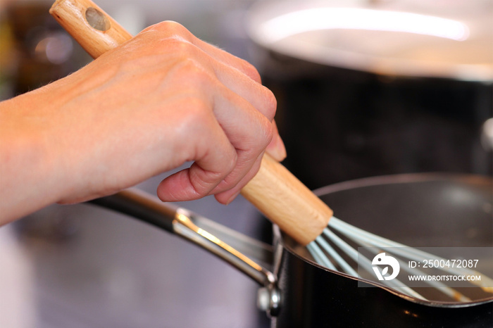 Woman’s hand holding a whisk, stirring sauce in a pan in the kitchen, close up with blurred backgrou