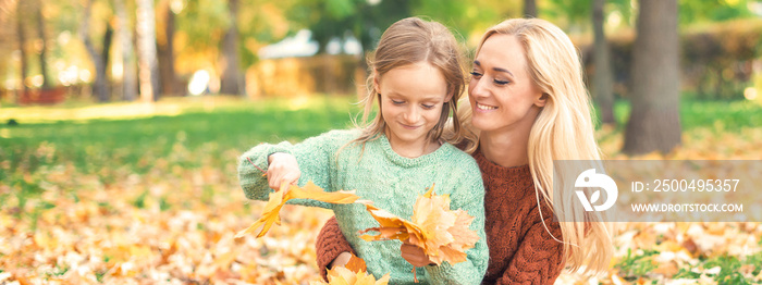 Happy young caucasian mother and little daughter holding autumn yellow leaves sitting at the park