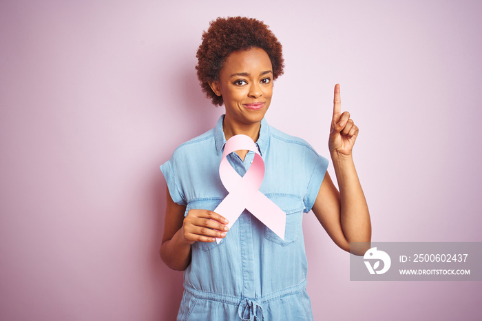 Young african american woman holding brest cancer ribbon over isolated pink background surprised wit