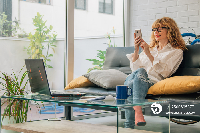 woman at home with mobile phone and computer sitting on the sofa