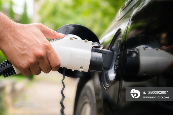 Mans hand inserting charger plug into electric car in green environment background. New energy vehicle, NEV is being loaded with electricity power. Ecology, modern day cars