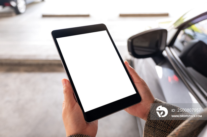 Mockup image of a woman holding and using digital tablet with blank white screen next to the car in parking lot