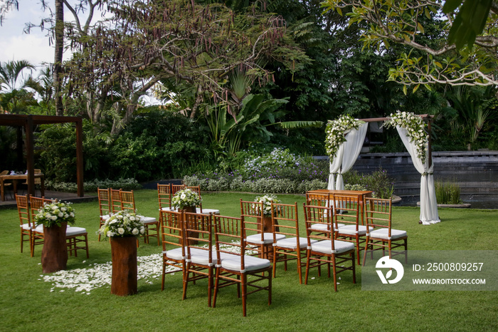 Beautiful romantic outdoor wedding setup in tropical garden, wooden chairs and simple gazebo decorated with white flowers