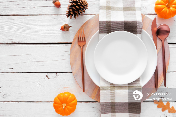 Autumn harvest or thanksgiving dinner table setting with plate, flatware, check print napkin, pumpkins and decor. Top view on a white wood background.