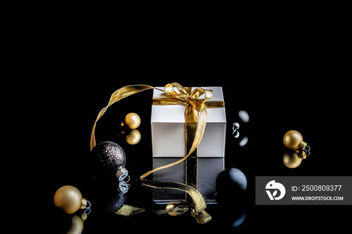 Christmas background golden. White gift box with golden ribbon, New Year balls and winter tree in xmas composition on black background for greeting card. Copy space. Winter holidays, New Year.