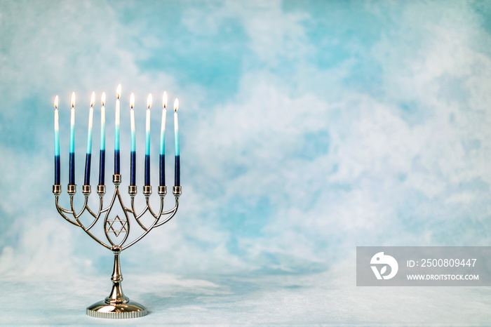 A silver menorah for the Jewish holiday Hanukkah with burning glowing eight candles on fire lit up on a blue background with copy space