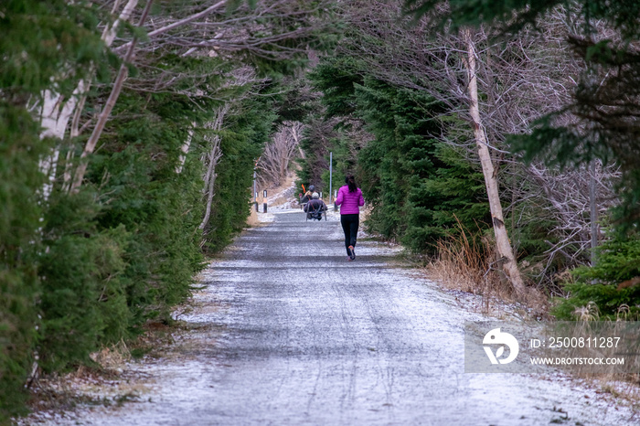 A female jogger runs along a trail in winter with trees enclosing like a tunnel the footpath. She is wearing a bright pink coat and black pants. There are two handcycles in front of the female jogger.