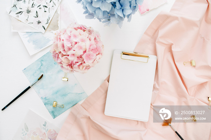 Watercolor artist home office desk workspace with copy space clipboard, pastel hydrangea flowers bouquet, watercolor painting, peachy blanket. Flat lay, top view.