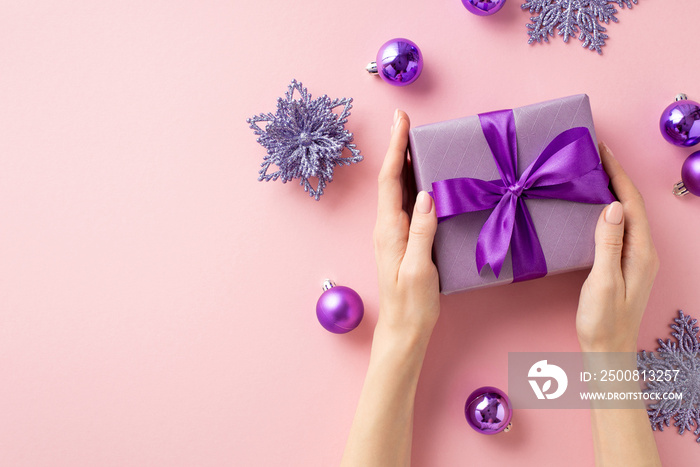 New Year concept. First person top view photo of woman’s hands holding violet giftbox with ribbon bow over purple baubles snowflake flower ornaments on isolated pastel pink background with copyspace