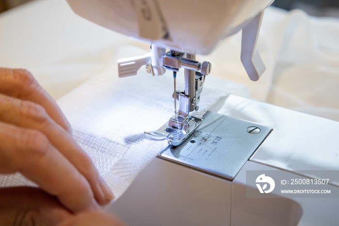 Cropped female hands sewing fabric in manufacturing machine at workplace. Needle pressure foot with blurred white background. Apparel production. Close up top side view. Selective focus copy space.