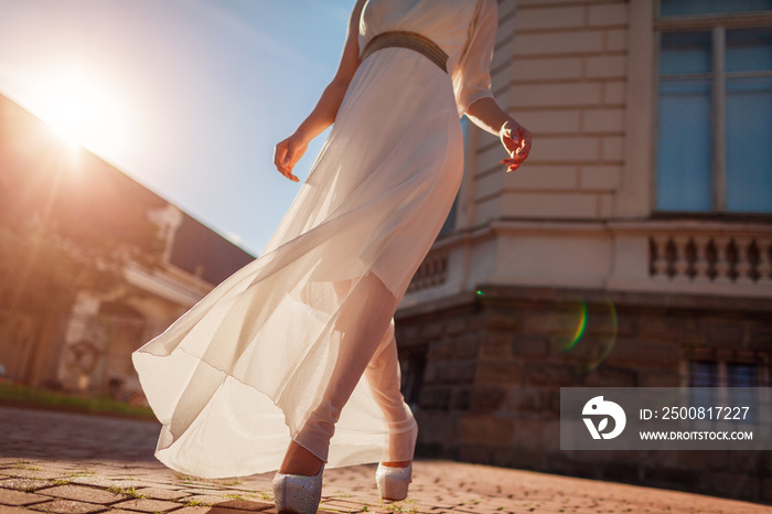 Beautiful woman wearing white wedding dress outdoors in summer. Lady walking by palace at sunset