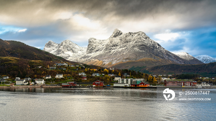 Ørnes in Nordland, Norwegen mit bunten Häusern, herbstlichen Wäldern und frisch verschneiten Bergen im Hintergrund. Interessante Wetterstimmung mit Gewitterfront.