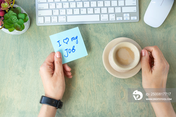 female hands hold note with words I love my job, cup of coffee, keyboard, cactus and mause on a wooden table. concept of good job. top view of the desktop.