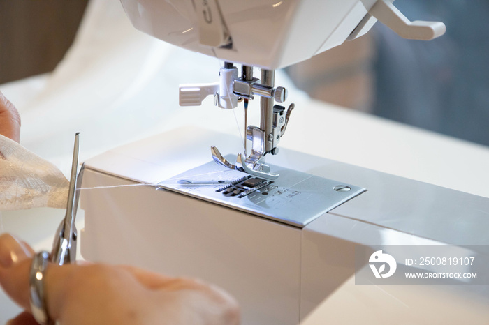 Close up female hands sewing fabric in manufacturing machine at workplace cutting thread with scissors. Tailor seamstress holding white textile for clothing apparel production. Close up top side view.