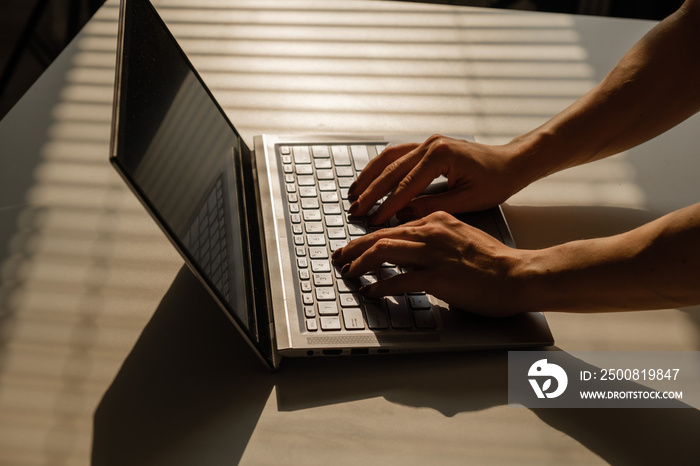 A woman is typing on a laptop keyboard on a white table. The shadow from the blinds falls on the desktop.