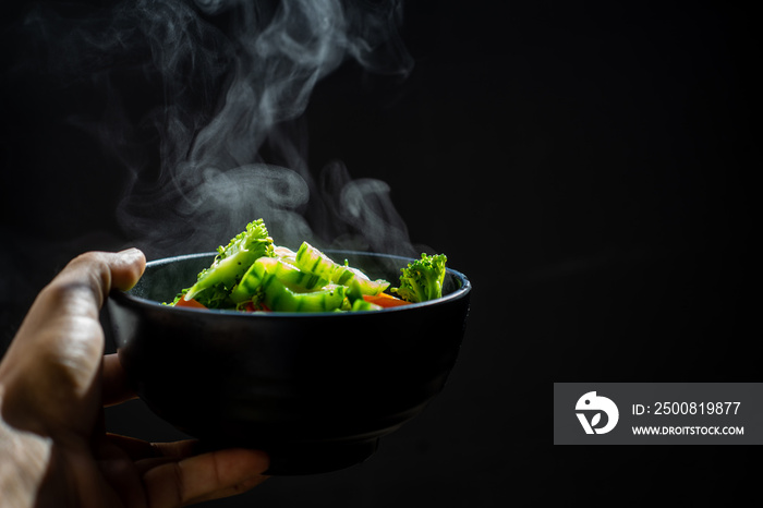 Female hands holding Mixed vegetables in bowl with steam and smoke on dark background selective focus.