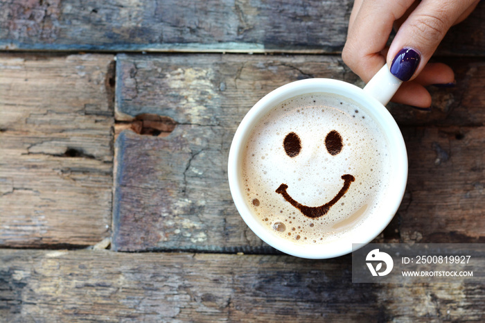 Happy face on cappuccino foam, woman hands holding one cappuccino cup on wooden table