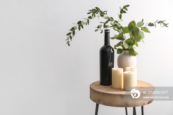 Beautiful burning candles with eucalyptus in vase and bottle of wine on table against light background