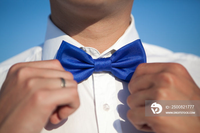 Groom in a white shirt straightens a blue bow tie against a blue sky