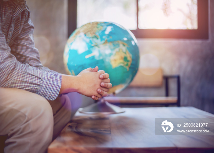 A young man sit on  wooden chair while praying to God over blurred world globe on wood table in door against window light,  great mission or world save concept with copy space.