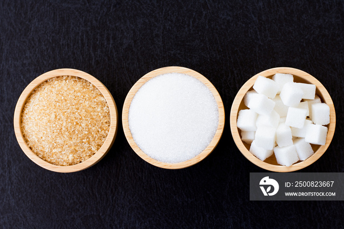 Three various types of sugar (brown granulated, white sand and sugar cubes ) in wooden bowl isolated on dark table background.