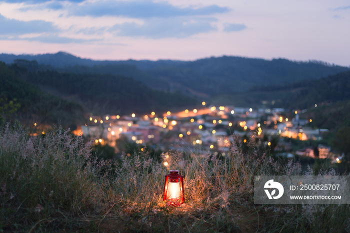 Kerosene lamp lit in the middle of the grass with the lights of a village in the background. Concept of light illuminating darkness or energy.