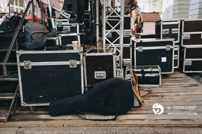 A black guitar case stands on a wooden flooring of the scene, next to boxes and music speakers. The concept of preparation for an outdoor concert.