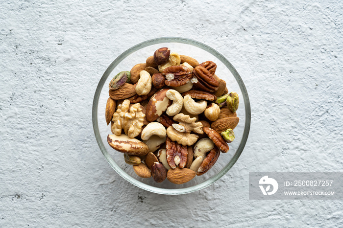 Viewed from above, against a textured concrete background, an isolated glass bowl holds a large variety of shelled nuts.