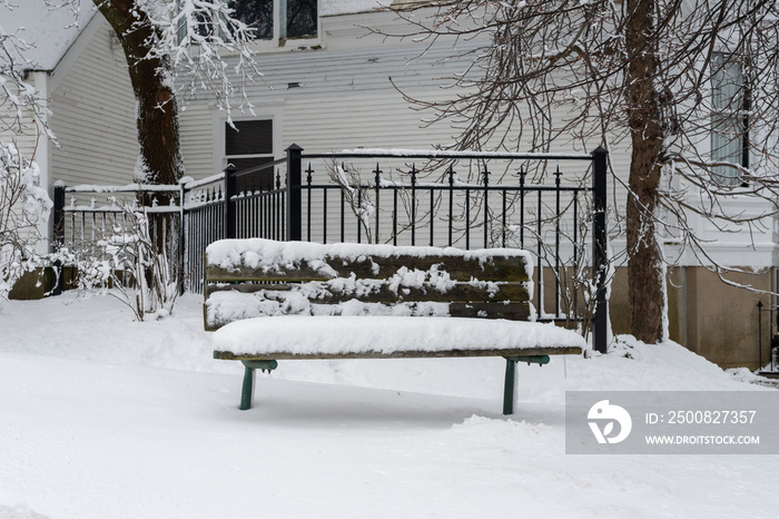 A wooden park bench on a street corner with a wrought iron fence and a white wooden building in the background. The seat is covered in fresh white fluffy snow and ice. The ground is snow covered.