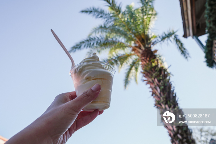 Hand holds up a cup of soft serve pineapple vanilla swirl ice cream, with palm trees in the background