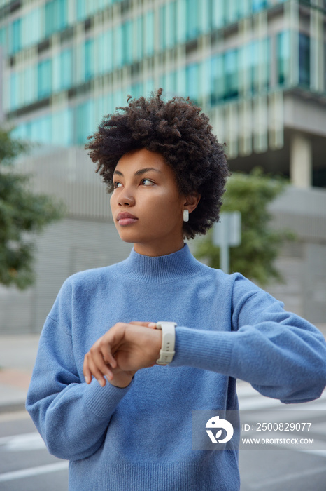 Outdoor shot of pensive curly haired Afro American woman waits for someone at street checks time onn wristwatch wears casual blue jumper strolls in downtown poses outside considers something