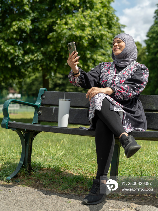 UK,Sutton,Woman in headscarf sitting on bench in park,talking on smart phone